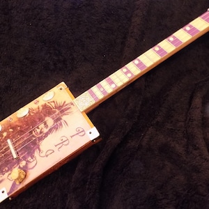 May include: A cigar box guitar featuring a rectangular wooden body with a printed graphic of a person and the text PRG. The neck has a light wood fretboard with purple fret markers and circular inlays. Metal hardware includes a bridge and tuners.