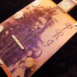 Op de afbeelding: Een sigarenkistgitaar met een paarse en beige grafische print van een persoon met dreadlocks. Het instrument bevat zilverkleurige metalen onderdelen, een cilindrische brug, decoratieve ovale accenten en de letters PRG op de klankkast.