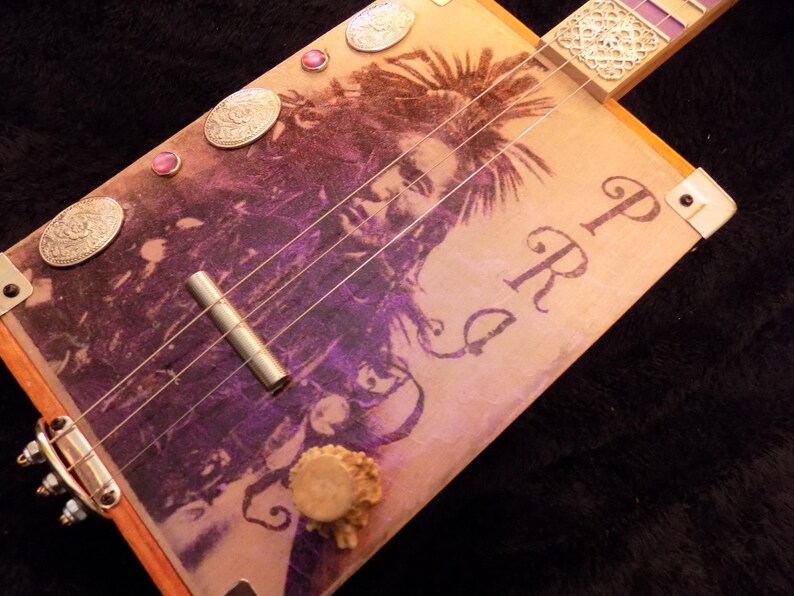 Op de afbeelding: Een sigarenkistgitaar met een paarse en beige grafische print van een persoon met dreadlocks. Het instrument bevat zilverkleurige metalen onderdelen, een cilindrische brug, decoratieve ovale accenten en de letters PRG op de klankkast.