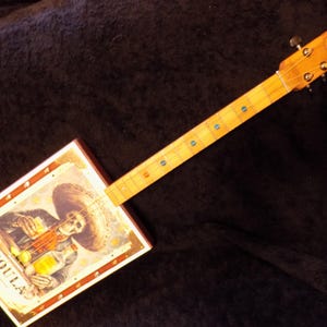 May include: A handmade cigar box guitar with a wooden neck and fretboard. The rectangular box features a colorful image of a skeleton with a sombrero, holding a bottle of tequila. The word "TEQUILA" is printed on the box.