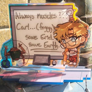 Puede incluir: Un expositor de acrílico holográfico con un personaje de dibujos animados de pelo castaño y gafas que sostiene un lápiz grande. El panel de fondo muestra el texto: Always Muscles?? Carl...(foggy) Save Erid. Save Earth. Se ven un pequeño robot y un globo terráqueo.