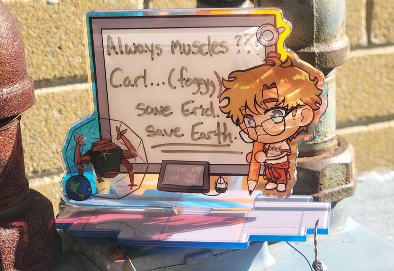 May include: A holographic acrylic standee featuring a cartoon character with brown hair holding a pencil, standing next to a whiteboard with the text: Always Muscles?? Carl...(foggy) save Erid. Save Earth. A small globe and robotic figure are visible.