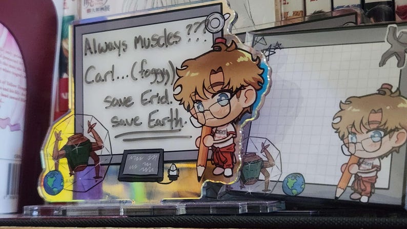 May include: An acrylic standee featuring a chibi character with brown hair and glasses holding a large pencil. The background panel displays a whiteboard with the text "Always muscles??? Carl...(foggy) save Erid. save Earth." and a small drawing of a planet.