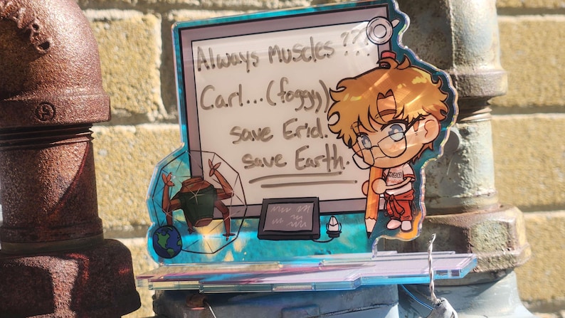 May include: A clear acrylic standee featuring a cartoon character with blonde hair holding a large pencil. The design includes a white board with the text Always Muscles ??? Carl... (foggy) Save Erid. Save Earth. and a small robot graphic.