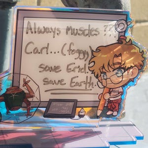 May include: An iridescent acrylic standee featuring a cartoon character with blonde hair and glasses holding a large pencil. The background panel displays the text: Always Muscles?? Carl...(foggy) save Erid. Save Earth. A small robotic figure and a tablet are also visible.
