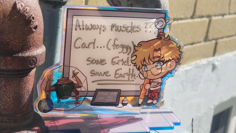 May include: An iridescent acrylic standee featuring a cartoon character with blonde hair and glasses holding a large pencil. The background panel displays the text: Always Muscles?? Carl...(foggy) save Erid. Save Earth. A small robotic figure and a tablet are also visible.