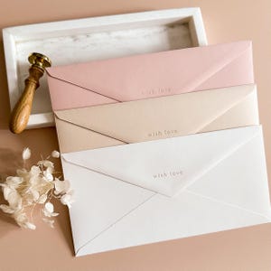 Könnte beinhalten: Drei pastellfarbene Umschläge, rosa, beige und weiß, sind in einem weißen Tablett gestapelt. Jeder Umschlag ist mit den Worten "with love" bedruckt. Ein Wachssiegel aus Holz und Gold sowie Trockenblumen sind ebenfalls im Bild.