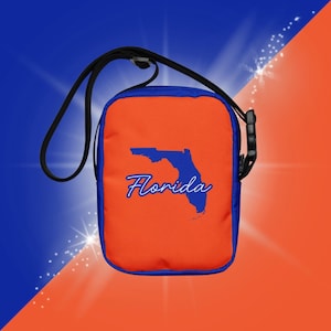 Op de afbeelding: Oranje crossbodytas met blauwe rand en riem. De tas heeft een blauwe omtrek van Florida met het woord "Florida" in script. De achtergrond is diagonaal verdeeld met blauw en oranje.