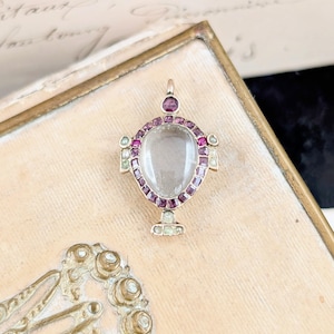 Peut inclure: Pendentif doré avec une pierre centrale transparente de forme ovale, entourée de petites pierres précieuses rouge foncé et vert pâle. Le pendentif présente un design décoratif et est posé sur un fond beige clair.