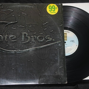 May include: A vinyl record album with a black cover featuring the band name "Cate Bros." in a stylized font. The cover has a textured, dark gray appearance with water droplet details. A yellow and green sticker is in the upper right corner.
