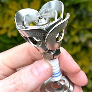 May include: A metallic sculpture shaped like a flower, constructed from a spark plug and curved metal washers. The center features a spark plug electrode, while the petals are formed from silver-colored washers. The text "motocepti" is visible.
