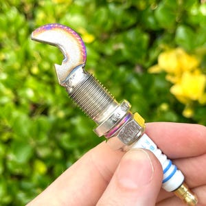 May include: A custom bottle opener made from a spark plug. The opener has a silver finish with iridescent accents on the curved top. The spark plug has a white ceramic insulator with blue stripes and a gold-colored tip.