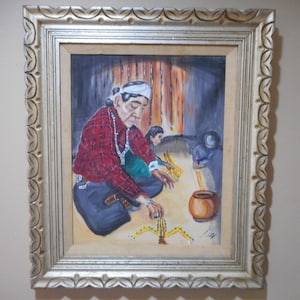 May include: A framed painting depicting three people, two women and a child, sitting on the floor and weaving with yellow and brown threads. The woman in the foreground is wearing a red and black plaid shirt and a white headband. The painting is set in a rustic interior with a wooden wall and a clay pot.