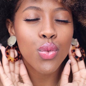 May include: A close-up of a person's face with their eyes closed and lips puckered. They are wearing a nose ring and large, round earrings with a tortoiseshell pattern.