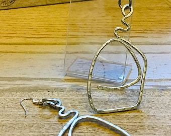 May include: Silver-toned, abstract dangle earrings. Each earring features a hammered, irregular geometric shape suspended from a curved wire. The design includes a looping, wavy element connecting the hook to the main shape. The earrings are displayed on a wooden surface.