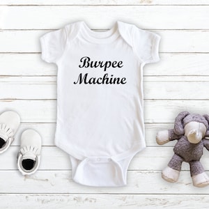 May include: White baby onesie with black text that reads "Burpee Machine". The onesie is on a white wooden background with a pair of white baby shoes and a gray stuffed elephant toy.