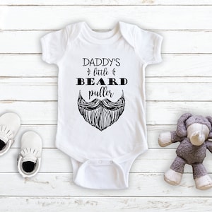 May include: White baby onesie with black text that reads "Daddy's little beard puller" and a black and white illustration of a beard.