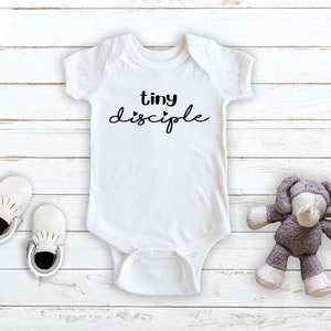 May include: White baby onesie with black text that reads "tiny disciple" with a small heart symbol before the word "disciple". The onesie is on a white wooden background with a pair of white baby shoes and a gray stuffed elephant toy.
