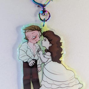 Może przedstawiać: Brelok z motywem Upiora w Operze. Brelok posiada kolorowe zapięcie w kształcie serca. Ozdoba przedstawia kreskówkową ilustrację Upiora i Christine, z Upiorem w białej koszuli i Christine w białej sukni.