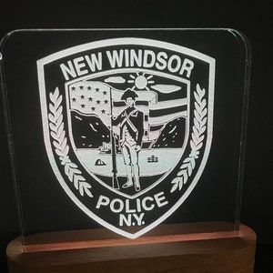 Könnte beinhalten: Ein transparentes Acryl-LED-Nachtlicht mit dem Emblem der New Windsor Police N.Y., auf einem Holzsockel. Inklusive Fernbedienung zur Farbeinstellung. Das Emblem zeigt einen Soldaten, eine Flagge und Text.