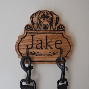May include: Wooden dog leash holder with two hooks. The holder features a laser-engraved image of a dog's head peeking over the top and the name "Jake" below.