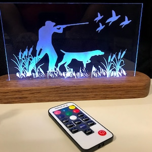 May include: A wooden base with a clear acrylic panel featuring a silhouette of a hunter with a dog and flying ducks. The panel is illuminated with blue LED lights and comes with a remote control to change the color of the lights.