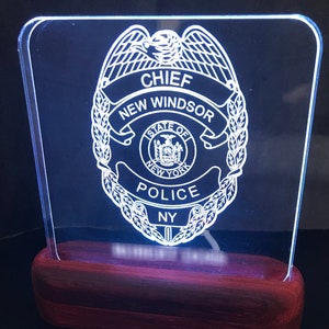 Könnte beinhalten: Eine weiße Acrylplatte mit einem Polizeibadge-Design. Das Abzeichen trägt die Aufschrift "Chief", "New Windsor", "State of New York", "Police" und "NY". Die Platte wird von hinten beleuchtet und steht auf einem dunklen Holzsockel.