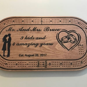 May include: A wooden cribbage board with a personalized inscription that reads "Mr. And Mrs. Bruce 3 kids and 4 amazing years Est. August 26, 2017". The board features a heart with two interlocking rings and a silhouette of a couple.