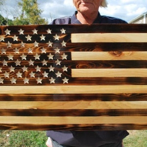 May include: A wooden American flag with a distressed, burnt look. The flag is made of multiple wood planks, with the stars and stripes painted in white. The flag is displayed against a green grassy background.