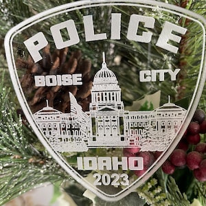 Puede incluir: Un adorno de plástico transparente con forma de placa de policía con las palabras "Police", "Boise City", "Idaho" y "2023" grabadas en él. El adorno presenta un dibujo lineal del edificio del Capitolio del Estado de Idaho.