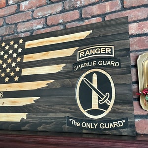 May include: A wooden American flag with a distressed look. The flag is partially painted black and has a gold-colored Ranger tab with the text "Ranger" and "Charlie Guard". Below the tab is a black and gold emblem with a sword and the text "The Only Guard".