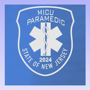 Può includere: Un distintivo bianco su sfondo blu. Il distintivo ha la forma di uno scudo e presenta una stella della vita bianca con un serpente avvolto attorno al bastone. Il testo sul distintivo recita "MICU PARAMEDIC STATE OF NEW JERSEY 2024".