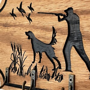 May include: Wooden key holder with a silhouette of a hunter with a dog and ducks flying overhead. The key holder has four hooks and is decorated with a wood grain finish.