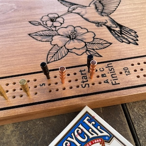 Puede incluir: Un tablero de juego de madera con un colibrí y un diseño floral. El tablero tiene una línea de inicio y final con los números 5 y 120. Hay clavijas en el tablero y una baraja de cartas de Bicycle.