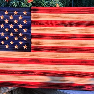 May include: A wooden American flag with red, white, and blue stripes. The stars are gold and the wood has a distressed look.