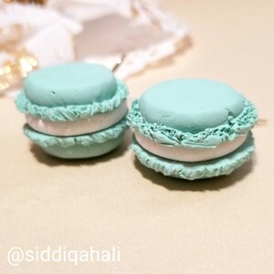 Puede incluir: Un par de pendientes de macarons de color azul turquesa con relleno blanco. Los macarons tienen una superficie texturizada y están hechos de arcilla polimérica.
