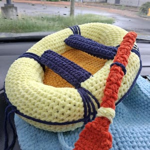 Pode incluir: Uma balsa de brinquedo em crochê com um anel inflável amarelo-pálido, um piso interior azul-escuro e dois bancos transversais azul-escuros. Um remo laranja em crochê está preso à lateral da balsa, que repousa sobre uma base azul-clara.