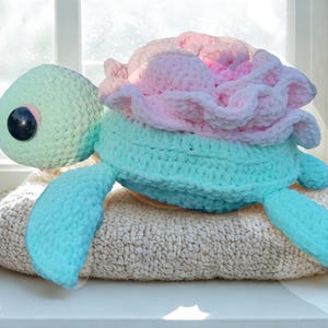 May include: A handmade crochet turtle in shades of mint green, light blue, and pink. The turtle has a large, ruffled flower design on its back and a large black eye. It is resting on a beige, textured surface.