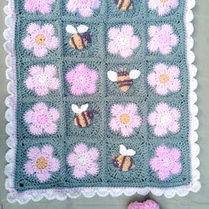 Könnte beinhalten: Handgehäkelte Decke mit einem sich wiederholenden Muster aus rosa Blumen und Hummeln auf grauem Hintergrund. Die Decke hat einen weißen, gewellten Rand und eine passende rosa Blume. Ideal für ein Baby oder Kind.