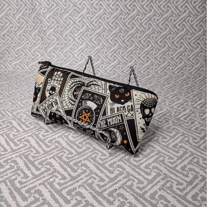 May include: A black and white zippered pouch with a repeating pattern of occult symbols, a black cat, and the text "BLACK CAT THE MOON". The pouch has a black zipper and is displayed on a decorative stand.