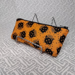 May include: An orange pencil case with a black cat face and star pattern. The zippered pouch is displayed on a silver wire stand. The cat faces have a crescent moon on their foreheads.