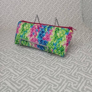 May include: A rectangular pencil case with a colorful scale pattern in shades of pink, blue, and green. The case has a red zipper and trim. The background is a light gray patterned surface.