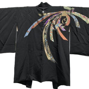 May include: A black kimono jacket featuring wide sleeves and a prominent decorative motif on the upper right side. The design consists of multiple flowing ribbons in various colors, including gold, blue, and red, with intricate floral patterns.