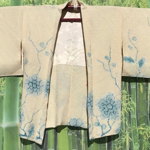 May include: Cream-colored kimono with blue floral embroidery. The open-front garment features large floral designs on the sleeves and lower front, with delicate branch and leaf patterns. The interior lining is a white, patterned fabric.
