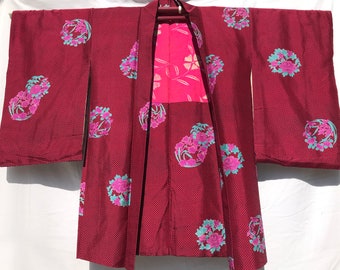 May include: A burgundy kimono with a repeating pattern of small black dots. The kimono features large floral designs in pink and teal. The interior panel is a bright pink with a floral design. The kimono is hanging on a wooden hanger.