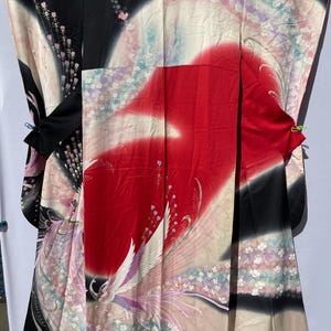 May include: A traditional Japanese kimono with a vibrant design. The kimono features a striking red central panel, contrasted by black and cream sections. Delicate floral and bird motifs in pink, white, and purple adorn the fabric, creating an elegant and artistic garment.
