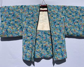 May include: A vintage kimono with a blue background and a repeating pattern of colorful fan-like shapes. The kimono has wide sleeves and a cream-colored lining. It is hanging on a wooden hanger.