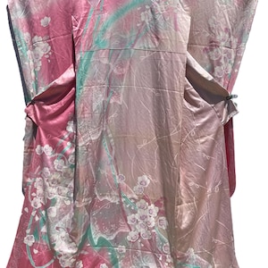 May include: A pink kimono featuring a satin-like finish with a repeating pattern of white cherry blossoms and flowing teal ribbons. The garment has wide sleeves, a high collar, and a long, flowing silhouette with a gradient color transition.