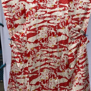 May include: A vintage kimono in cream and red, featuring a flowing pattern of stylized waves and floral accents. The fabric has a subtle sheen, and the design includes small yellow flower clusters.