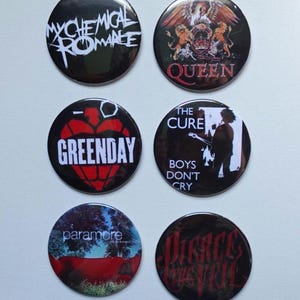 May include: A set of six circular pin-back buttons featuring various band logos and imagery. The designs include My Chemical Romance, Queen, Green Day, The Cure with the text Boys Don't Cry, Paramore, and Pierce the Veil on black and colorful backgrounds.